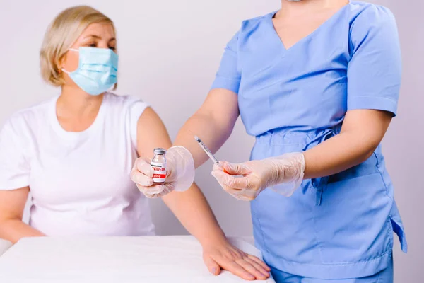 Fotografía recortada. Un médico con guantes de látex desechables sostiene una ampolla con la vacuna covid 19 y una jeringa a la cámara. Paciente femenina caucásica mayor en el fondo con una máscara médica. — Foto de Stock