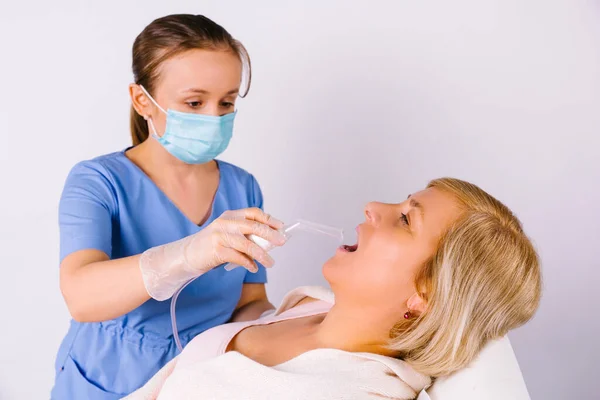 Νεαρός γιατρός με ιατρική μάσκα και γάντια λατέξ αντιμετωπίζει το λαιμό μιας μεγαλύτερης γυναίκας με νεφελοποιητή σε λευκό φόντο. Καταπολέμηση του ιού κρυολογήματα. — Φωτογραφία Αρχείου