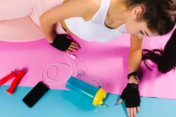 Giovane sportiva su tappetino yoga rosa con manubri, telefono e cuffie. Concetto home fitness. Foto dall'alto. — Foto Stock