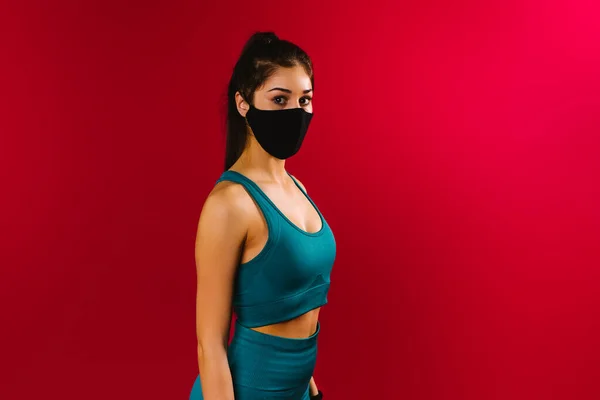 Porträt eines Sportlers in Top und Leggings auf rotem Hintergrund mit einer schwarzen Schutzmaske. Das Konzept der Ausbildung während der Quarantäne. — Stockfoto