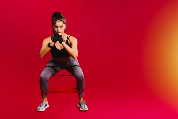 Kafkasyalı pompalı kız kırmızı arka planda yan tarafları olan elastik bir bantla bacak kaslarını sallıyor..