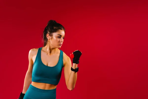 A brunette girl with dumbbells looks to the side and shakes the muscles. Red background with side space. — Stock fotografie