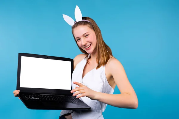 Ragazza bionda con le orecchie da coniglietto bianche che tiene un quaderno, mostra l'indice allo spazio vuoto bianco, lo schermo del taccuino mockup. Sorridi e guarda la fotocamera, su sfondo blu con spazio laterale — Foto Stock