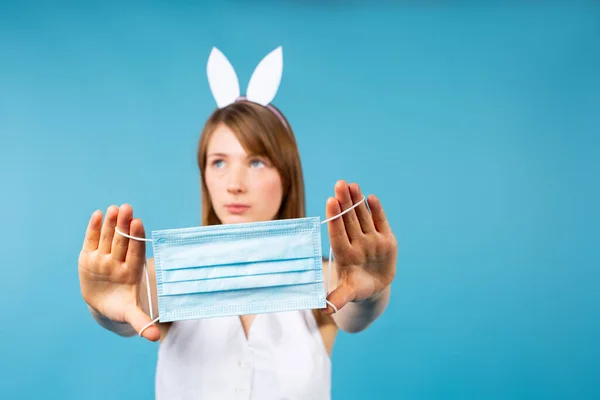 En ung blond kvinna bär vita öron av en påskhare, visa en engångs medicinsk mask framför kameran, på en isolerad blå bakgrund med tomt utrymme för din annons — Stockfoto