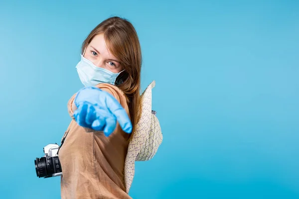 Menina turística pandêmica em máscara médica e luvas com chapéu de verão e câmera fotográfica isolada no fundo azul. Jornalista loira viajando. De pé com as mãos estendidas para saudação — Fotografia de Stock