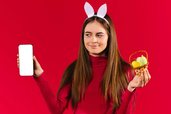 Кавказская девушка с кроличьими ушами смартфон и декоративные пасхальные яйца на красном фоне. — стоковое фото