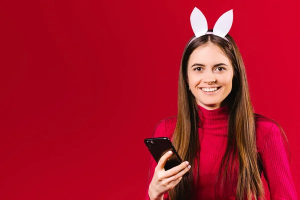 Счастливая молодая девушка блоггер с ушами и красивой улыбкой держит в руках смартфон и смотрит в камеру на красном фоне. — стоковое фото