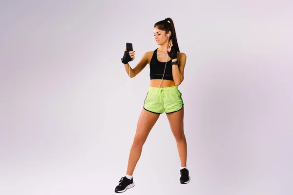 Studio foto van een jong sportmeisje met koptelefoon poseren en het nemen van een selfie op een smartphone. Witte achtergrond en lege kant advertentieruimte. — Stockfoto