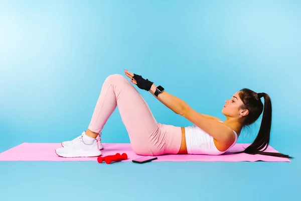 Esercizio sul tappeto fitness rosa. La ragazza bruna scuote la stampa. Sfondo blu con ritorno spazio vuoto. — Foto Stock