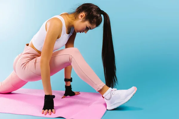 Uitgebreide foto van een brunette tijdens fitnesstraining. Uitrekken op het roze sporttapijt. Blauwe achtergrond met zijruimte. — Stockfoto