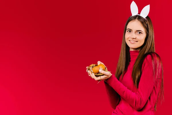 Симпатичная девушка с декоративными кроличьими ушами держит красочные пасхальные яйца на красном фоне с пустым боковым пространством. — стоковое фото