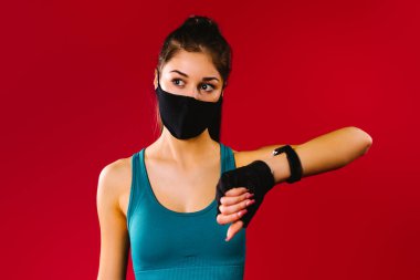Kırmızı arka planda bir fitness saati olan koruyucu siyah maskeli atletik kız yana bakıyor. Spor salgını konsepti