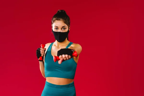 身穿防护面具的白人女孩把红色哑铃伸向摄像机。红色背景和侧面广告空间。大流行病期间的锻炼. — 图库照片