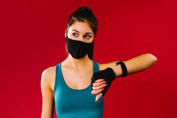 Sportowa dziewczyna w czarnej masce ochronnej z zegarkiem fitness na czerwonym tle wygląda z boku. Koncepcja pandemii sportu — Zdjęcie stockowe