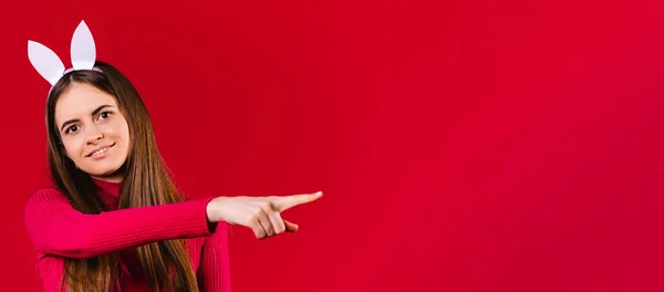 Banner, formato longo. Menina caucasiana com orelhas de coelho sorrindo olha para a câmera e aponta com o dedo indicador para um espaço vazio onde sua propaganda pode ser. Fundo vermelho. — Fotografia de Stock