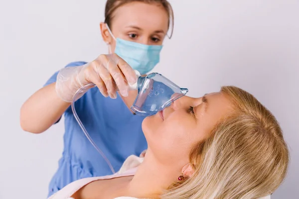 Flicka läkare i en medicinsk uniform och i en mask håller en nebulisator till en äldre kvinna på en vit bakgrund. Behandling av förkylningar. Förstorat foto. — Stockfoto