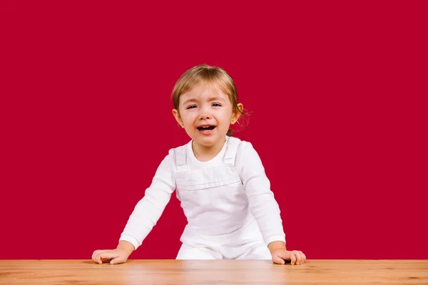 Rubia niña pequeña con ropa blanca casual está asustada y llorando niña de pie sobre la mesa de madera sobre fondo rojo — Foto de Stock