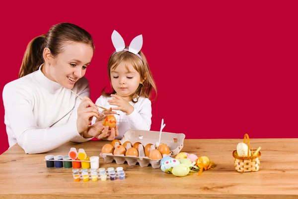 Portrait d'une jeune femme heureuse peignant des œufs de Pâques avec sa petite fille adorable faisant des décorations pour les vacances de Pâques. Fond rouge avec espace latéral — Photo