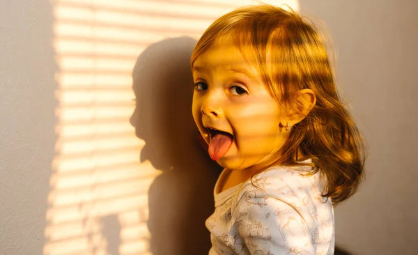 Niña aislada durante una pandemia en casa posando y mostrando la lengua a la cámara sobre el fondo de la luz del sol. — Foto de Stock