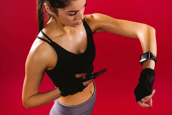 Mujer fitness cansada sudando tomando un descanso en el teléfono después de un entrenamiento difícil, rastreando el tiempo usando su reloj del brazo. Mujer joven escuchando música en la aplicación de teléfono inteligente para la motivación fitness —  Fotos de Stock