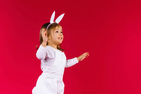 Vit litelblond flicka med vita kaninöron hoppar på röd bakgrund, tittar upp på tomt utrymme. Väntar överraskning på Happy påsk rabatt och försäljning — Stockfoto