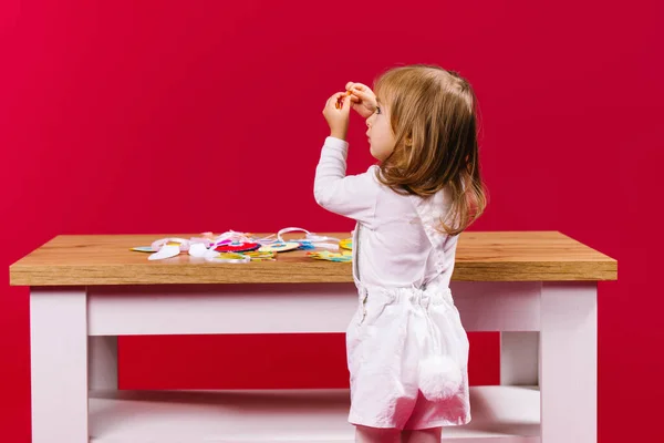 Foto trasera de rubia caucásica, niña en traje de Pascua conejito mostrando un detalle con las manos arriba, de pie cerca de la mesa de madera de huevos de papel. Colorida actividad hecha a mano en casa — Foto de Stock