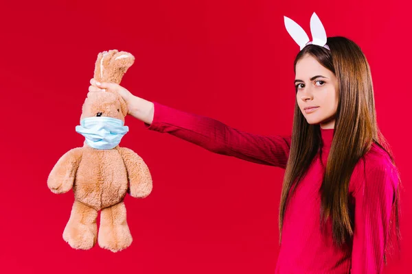 Morena séria jovem olhando para a câmera. Menina bonita segurando orelhas de coelho de Páscoa, pelúcia coelho de Páscoa com máscara médica em um fundo vermelho. Símbolo de Páscoa e férias pandêmicas — Fotografia de Stock