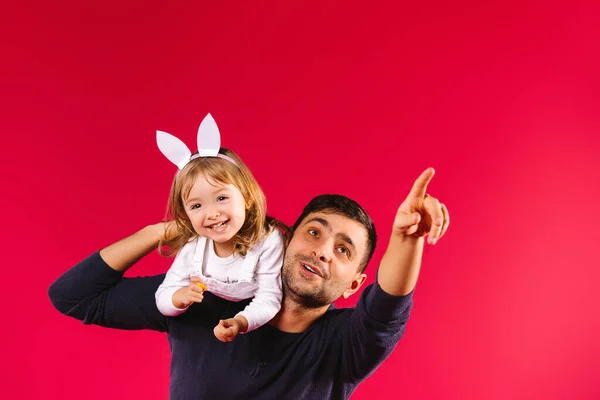 Kaukasiska vackra far bär sin lilla dotter på axeln. Man pekar pekfinger i avstånd på röd bakgrund med sidoutrymme, liten kvinnlig unge leende tittar på kameran — Stockfoto