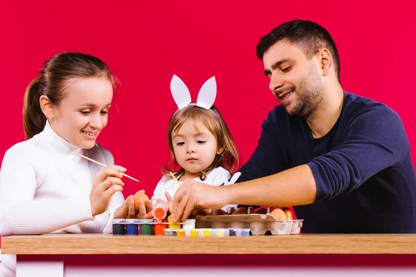 Une jeune famille caucasienne avec une petite fille, un lapin, se préparant pour les vacances et peignant des œufs de Pâques ensemble sur un fond rouge. Concept de Pâques en famille. — Photo
