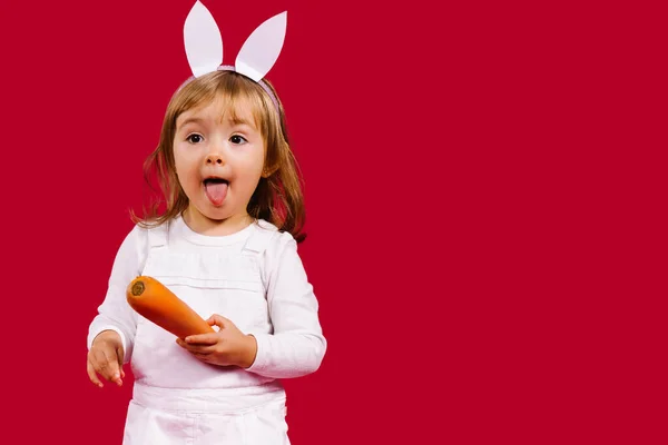 Påsk kanin flicka med en morot visar sin tunga ut och tittar åt sidan på en röd bakgrund med sidoutrymme. — Stockfoto