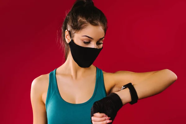 Ein Porträt einer Brünetten mit einer schwarzen Schutzmaske betrachtet ihre Errungenschaften auf einer Fitness-Uhr auf rotem Hintergrund.. — Stockfoto