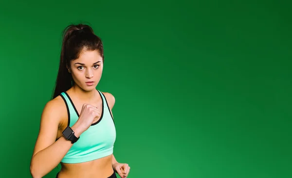 Young caucasian girl runner in light green sport top with a fitness watch on a green background looks at the camera while jogging. — Stock Photo, Image