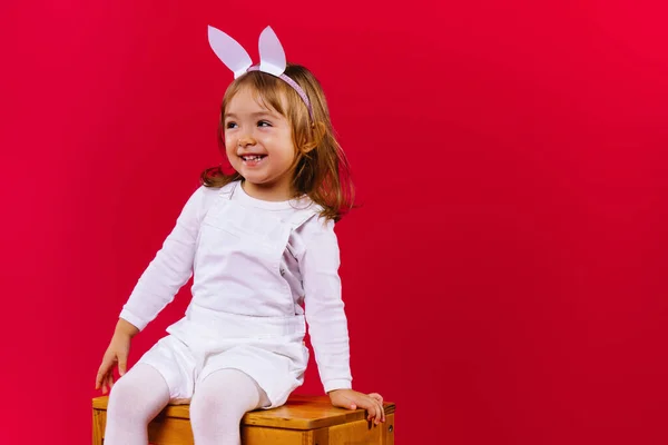 Glad liten flicka med kaninöron och i en vit kostym sitter på en stol på en röd bakgrund. Glada barn under påskhelgen. Sidoutrymme för reklam. — Stockfoto