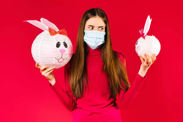 Férias de Páscoa durante uma pandemia. Menina morena em uma máscara médica segura coelhos decorativos em suas mãos em um fundo vermelho. — Fotografia de Stock