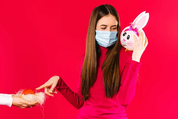 Uma menina com uma máscara médica segura um coelho decorativo em seu ombro e aponta com o dedo indicador para ovos de Páscoa pintados. Páscoa durante a quarentena. Fique em casa. — Fotografia de Stock