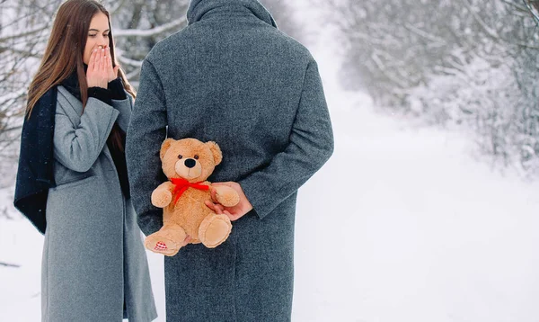 Κοντινό πλάνο δώρου, αρκουδάκι με κόκκινη κορδέλα. Νεαρός Καυκάσιος που κρατιέται πίσω από την πλάτη του για να κάνει έκπληξη στην κοπέλα του. Εκπληκτικό μελαχρινό δώρο αναμονής. Λευκό χιονισμένο φόντο πλευρά για τη διαφήμισή σας — Φωτογραφία Αρχείου