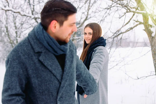 Felice giovane coppia caucasica che si tiene per mano e si guarda l'un l'altro, sorridente carino, mentre cammina nella foresta invernale, bianco sfondo innevato — Foto Stock