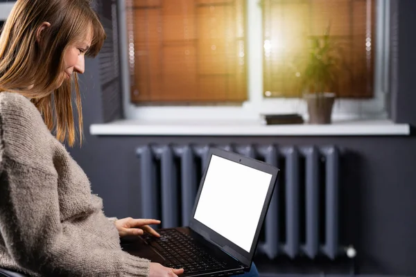 Ritratto di donna caucasica che usa il suo computer portatile a schermo bianco mentre si siede a casa alla luce del tramonto. Donna bionda si siede in casa a lavorare al computer con schermo bianco. Ministero degli Interni — Foto Stock