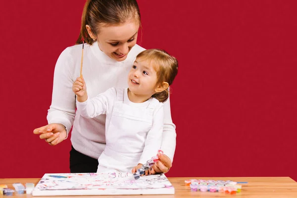 Mammas och dotters tidsfördriv hemma under karantänen. Vit kvinna med liten dotter färg med akryl på röd bakgrund med sido utrymme. — Stockfoto