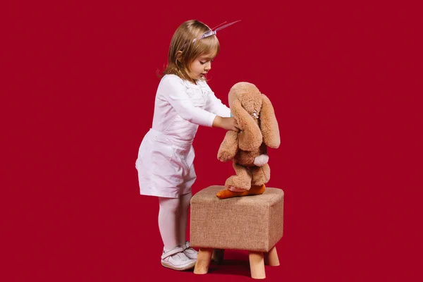 Niña del jardín de infantes caucásico se juega con un juguete suave sobre un fondo rojo. La chica se sienta un conejito suave en un taburete. Espacio en blanco para tu anuncio. — Foto de Stock