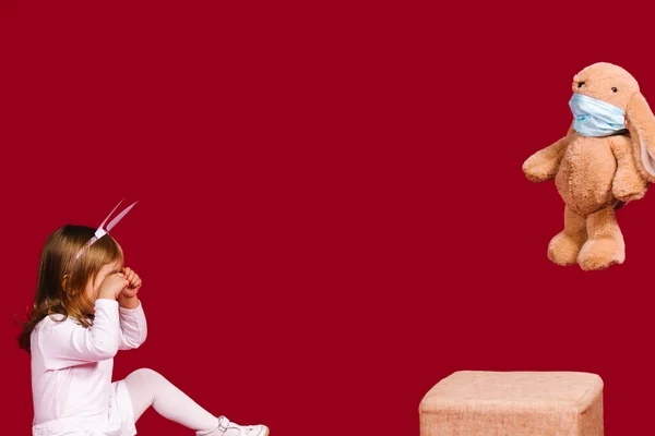 Una niña pequeña se sienta en el suelo llorando después de ver un juguete de conejo de Pascua con una máscara médica. Vacaciones de Pascua durante la pandemia covid 19. Los niños están rostroen. Fondo rojo y espacio vacío. — Foto de Stock