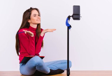 Genç esmer kadın, selfie çubuğuyla tutulan bir akıllı telefonun önünde. Kız gülümsüyor ve beyaz bir arka plan üzerine blog yazıyor..