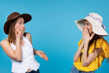 Mavi arka planda birbirine bakan iki bayan arkadaşı şok etti. Reklam metnin için ortası boş. Yazlık giysiler ve şapkalar giyen beyaz kadınlar. Satış ve promosyon