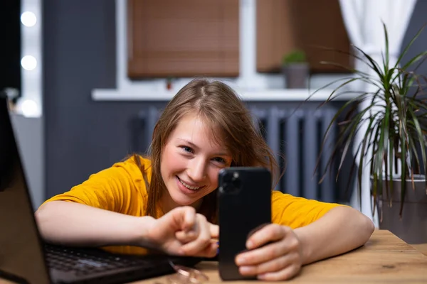 Giovane bionda felice con uno smartphone e un computer portatile accanto al tavolo. Lavora online a casa. — Foto Stock