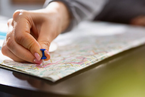 Female hand holding pushpin showing the location of a destination point on a map. Travel destination, pin on the map. Selective focus. Blue pushpin, map on table. High quality photo