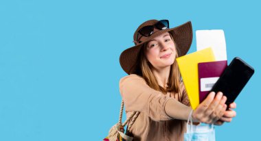 Banner, uzun format. Genç bir sarışın yaz şapkasıyla seyahat etmeye hazır. Bir cep telefonu, pasaport, uçak biletleri ve aşı hakkında sarı bir sağlık kartı, mavi arka plan. Yüksek kalite fotoğraf