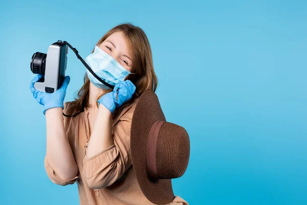 Блондинка-фотограф у медичній масці тримає літній капелюх на плечі і камеру в руці. Синій фон і бічний простір . — стокове фото