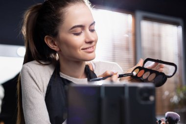 Elinde palet olan yakışıklı, beyaz tenli bir esmer makyaj yapıyor ve video blogu yazıyor. Online eğitim. Büyütülmüş fotoğraf. Yüksek kalite fotoğraf