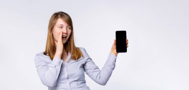Sancak- uzun format. Heyecanlı bir kadın çığlık atıyor, cep telefonunu boş siyah ekranlı, gri arka planda izole bir şekilde tutuyor. İnsanlar, yaşam tarzı, reklam. Kopyalama alanı oluşturun. Yüksek kalite fotoğraf