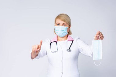 Tıbbi önlüklü bir doktor, koruyucu tıbbi maskeli bir kadın, dikkat çekmek için işaret parmağı gösteren, tıbbi bir maske tutan. Gri arka plan ve sağlık hizmetleri. Yüksek kalite fotoğraf